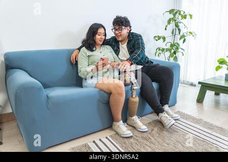 Diverse young couple with prosthetic leg relaxing on sofa, watching smartphone together Stock Photo