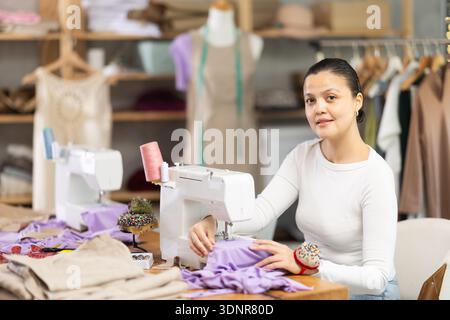 Attentive woman tailor sewing textile using sewing machine sitting at workplace in fashionable sewing atelier Stock Photo