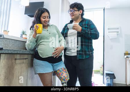 Pregnant woman with prosthetic leg embracing her baby bump beside her partner in the kitchen Stock Photo