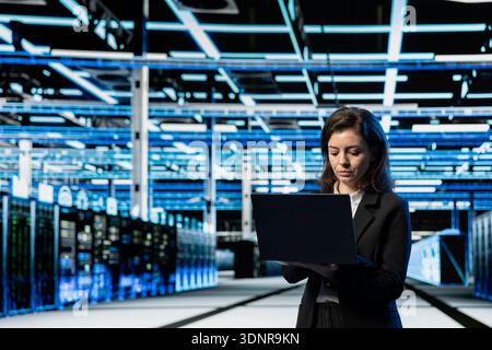 Data center engineer using laptop to do maintenance on cloud computing gear, VFX compositing overlay. Server farm worker walking between blue wireframe rackmounts using notebook Stock Photo