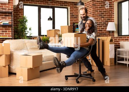 Happy black woman holding box while being pushed on chair by caucasian boyfriend in new home. Joyful multiethnic couple laughing and playing during moving day, excited to start new chapter together. Stock Photo