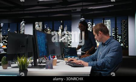 Tech agency system administrator using AI to optimize software performance in office late at night. IT expert at work using artificial intelligence to analyze code and do debugging Stock Photo