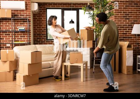 Young diverse couple standing in modern living room, carrying cardboard box and potted plant during apartment relocation. Happy multiethnic partners moving belongings in new home together. Stock Photo