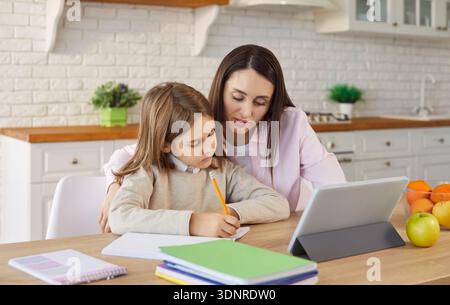 Mother, teacher and pupil daughter learning activity online using tablet computer, sitting together Stock Photo