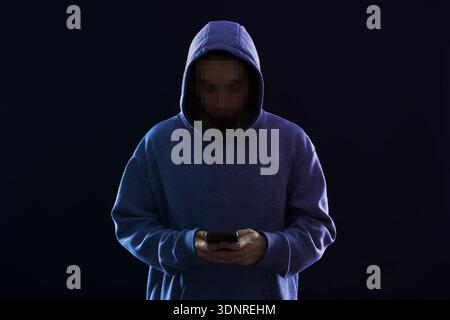 Criminal in hoodie using mobile phone on black background. Man with unrecognizable pixelated face Stock Photo