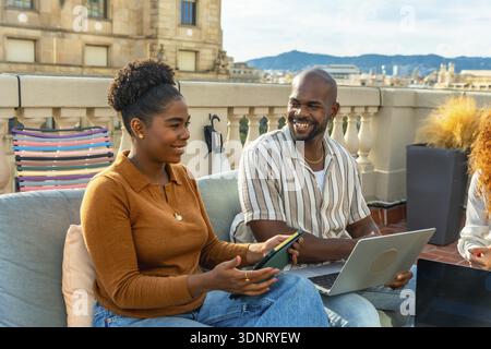 Two diverse colleagues working together and discussing ideas during a casual rooftop meeting, collaborating outdoors on a sunny day with city building Stock Photo
