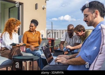Group of diverse business people using laptops and tablets while working together on a collaborative project during a casual meeting on an urban rooft Stock Photo