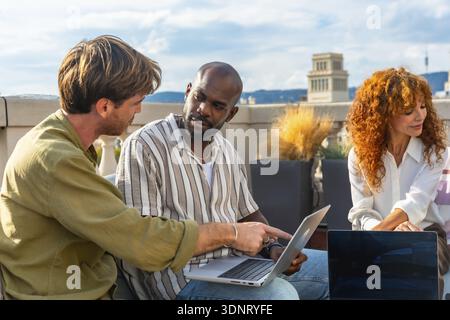 Diverse business professionals collaborating on a laptop during an informal rooftop meeting, sharing ideas and planning strategy in a sunny urban outd Stock Photo