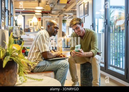 Two diverse male colleagues are sharing information on a mobile phone and laptop while working together, fostering teamwork and professional communica Stock Photo