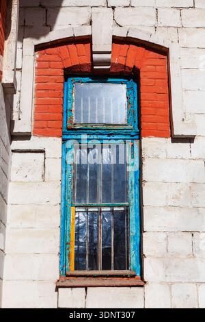 Weathered blue framed window set in brick arch and white stone wall, with textured glass panes. Aged details and contrasting materials suggest vintage Stock Photo