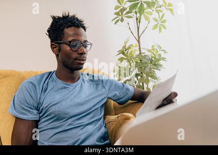 Young black man sitting on sofa working on laptop at home while checking paperwork, focused on online banking, bills and personal finance management. Stock Photo