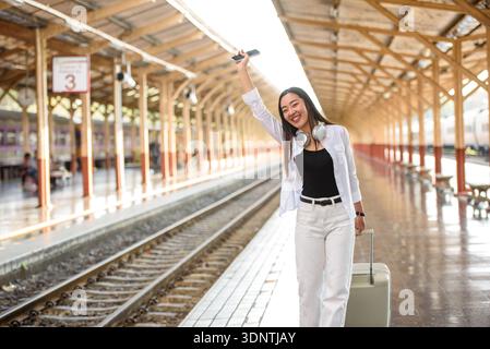Smiling young Asian woman waving while walking on a railway station platform, enjoying solo travel and freedom, modern lifestyle Stock Photo