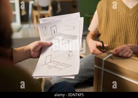Caucasian young adult men assembling furniture, one holding instruction manual with assembly diagrams while other using scissors to open cardboard box, working together indoors Stock Photo