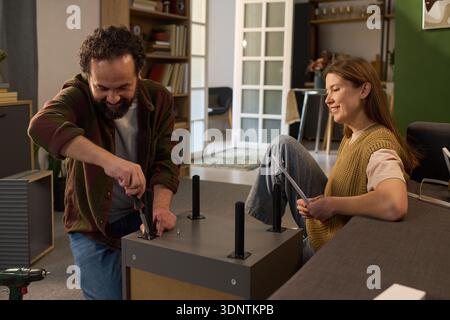 Caucasian middle aged man assembling furniture using screwdriver while Caucasian young adult woman sitting nearby holding instruction manual and smiling in modern home interior Stock Photo