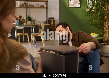 Caucasian middle aged man assembling furniture while sitting on floor, focusing on aligning parts, with Caucasian young adult woman observing in modern home interior Stock Photo