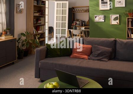 Modern living room featuring gray sofa with colorful pillows, open laptop on green coffee table, visible kitchen and dining area in background, indoor plants decorating space Stock Photo