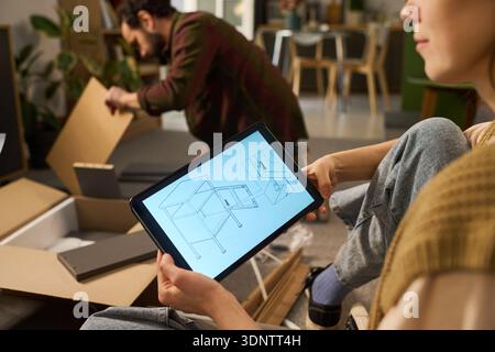 Caucasian young adult woman holding tablet displaying furniture assembly instructions while Caucasian young adult man assembling furniture in modern living room setting Stock Photo