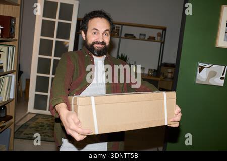 Portrait of Caucasian middle aged man smiling while holding cardboard box with both hands, standing indoors in modern home setting, looking directly at camera with cheerful expression Stock Photo