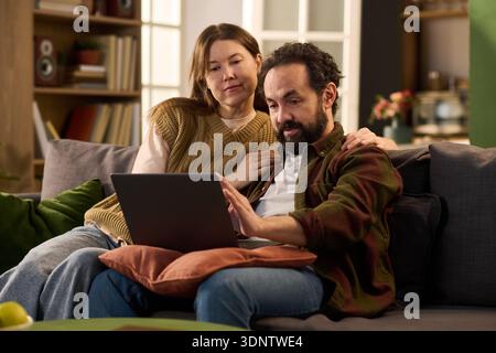 Caucasian middle aged man sitting on sofa using laptop with Caucasian middle aged woman embracing him from behind, both looking at computer screen and relaxing together at home Stock Photo