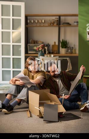 Vertical shot of Caucasian young adult woman and Caucasian young adult man sitting on floor unpacking cardboard box and examining instruction sheets together, in modern home setting Stock Photo