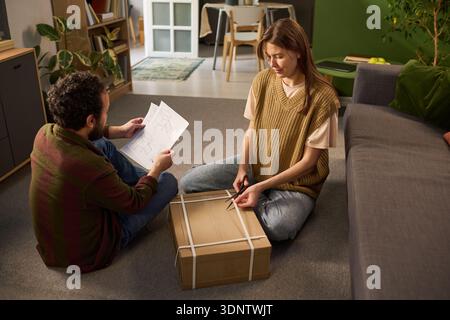 Young Caucasian woman sitting on floor opening cardboard box with scissors while young Caucasian man sitting nearby holding instruction papers, both preparing for assembly in living room Stock Photo