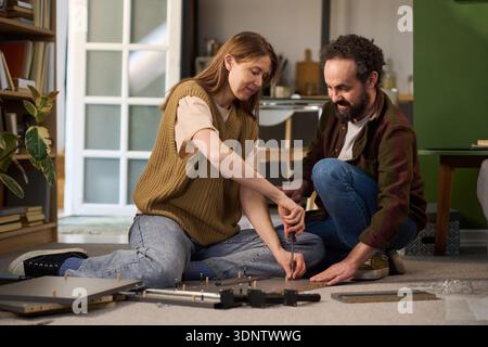 Caucasian young adult woman and Caucasian young adult man assembling furniture together on floor, using screwdriver and working as team, focusing on construction process in home setting Stock Photo