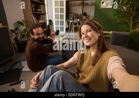 POV of young woman smiling and taking selfie while sitting on floor, Caucasian young man kneeling behind her holding camera and looking at her, both assembling furniture in modern living room Stock Photo