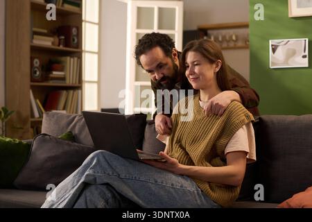 Caucasian young adult man standing behind Caucasian young adult woman sitting on sofa using laptop, both smiling and looking at computer screen in modern living room setting Stock Photo