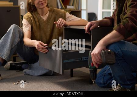 Caucasian young woman and Caucasian young man assembling furniture together on floor, woman holding drawer while man using electric drill, small hardware parts scattered nearby Stock Photo