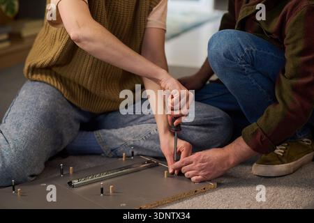 Caucasian young adult woman assembling furniture using screwdriver while kneeling on floor next to Caucasian young adult man assisting with hands holding wooden board Stock Photo