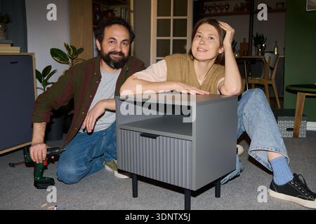 Portrait of Caucasian middle aged man kneeling with electric drill next to Caucasian middle aged woman sitting on floor assembling gray nightstand, both smiling and looking content Stock Photo