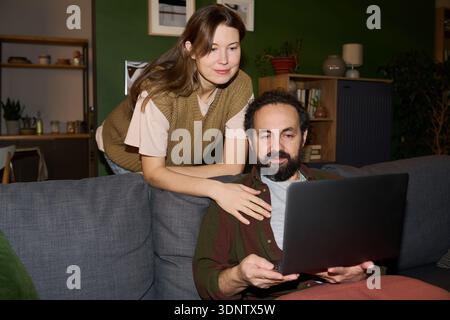 Caucasian young adult woman standing behind Caucasian middle aged man sitting on sofa using laptop together, both looking at computer screen, relaxed home environment visible Stock Photo