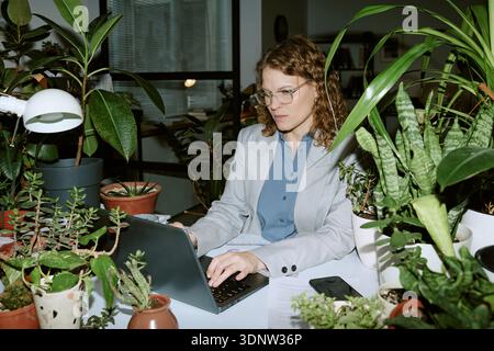 Young Woman Working Remotely on Laptop in Office Surrounded by Plants Stock Photo
