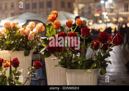 A close-up view of fresh red, orange, and peach roses in ornate white vases, glowing under warm street lights, evoking urban romance and special occas Stock Photo