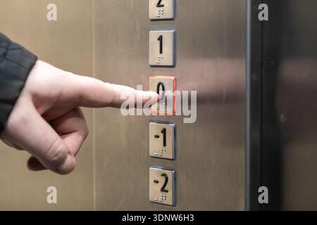Focus on the illuminated 0 button on a contemporary brushed steel elevator panel being pressed by a finger, showing easy access for all users Stock Photo