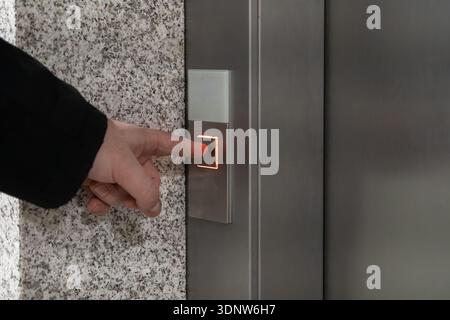 Close-up of a human hand activating a glowing call button on a sleek metallic elevator control panel, representing advanced technology and smooth vert Stock Photo