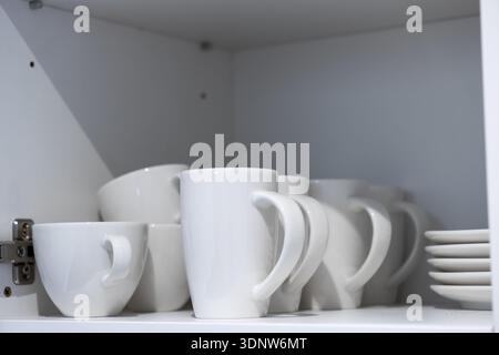 White ceramic mugs and plates neatly arranged on a shelf inside a kitchen cupboard, emphasizing efficient home organization and daily essential use fo Stock Photo