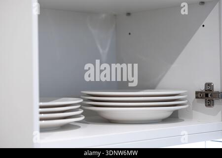 Close-up of bright white ceramic plates stacked in neat piles inside a modern, organized kitchen cabinet, emphasizing essential, practical home storag Stock Photo