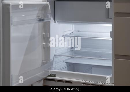 Brightly lit interior of a modern open white empty refrigerator showing clean glass shelves and door compartments, representing potential hunger, pove Stock Photo