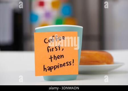 close view of an orange sticky note reading coffee first then happiness attached to a mug, with a plain bagel on a plate behind it in a home kitchen Stock Photo