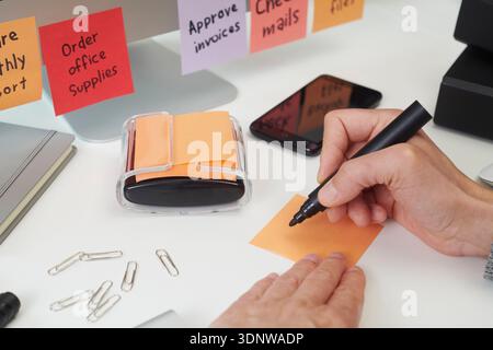 closeup view of a man about to write with a black marker on a blank orange sticky note on a clean office workspace, surrounded by stationery, and colo Stock Photo