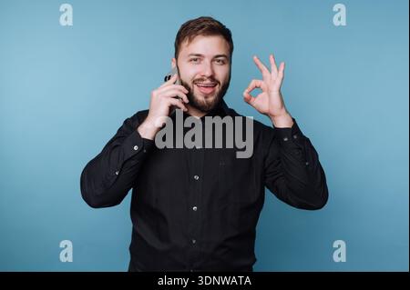 A cheerful young man stands confidently against a bright blue background. He is talking on his phone with excitement, making an okay gesture with his Stock Photo
