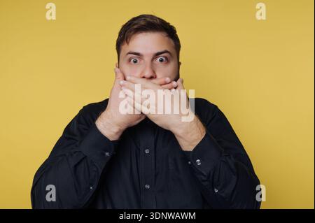 A young man stands stunned, hands covering his mouth in shock. His wide eyes and vivid black shirt contrast sharply with the vibrant yellow background Stock Photo