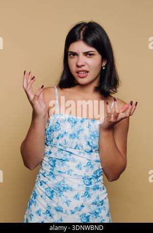 A young woman with dark hair showcases a puzzled expression while wearing a light blue floral dress. Her hands are raised in confusion, emphasizing he Stock Photo
