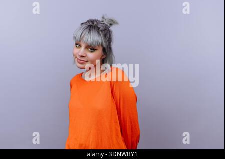 A cheerful young woman stands confidently, wearing an eye-catching orange sweater. Her playful silver hair complements her charming smile, creating a Stock Photo