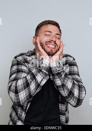 A young man radiates joy as he smiles broadly, hands on his cheeks, showcasing his delight in the cheerful atmosphere of a comfortable indoor space. Stock Photo