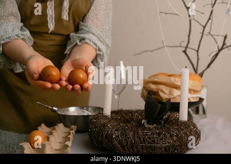 Woman painting eggs and preparing the Easter table, arranging candles and dishes with care and warmth at home. Stock Photo