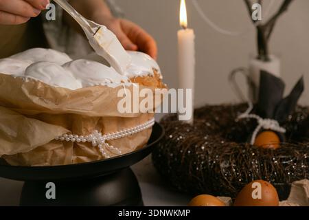 In cozy kitchen, she spreads icing on Easter cake, with eggs, candles, twig wreath on table, and beaded branch in background. Stock Photo