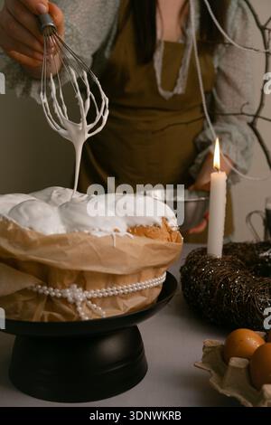 Woman brushes white icing on Easter cake, table decorated with eggs, candles, twig wreath, and beaded branch in background. Stock Photo