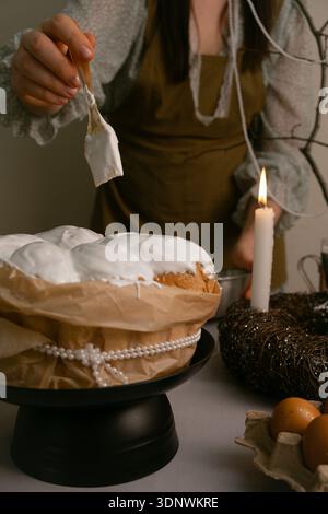 Woman carefully spreads white icing on Easter cake on a festively decorated table for Holy Easter celebration. Stock Photo